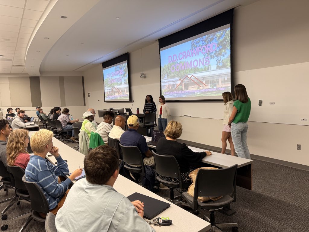 Class presentation for Crawford Elementary, a group of four students stand by a whiteboard and present work to a crowd seated in front of them at worktables.