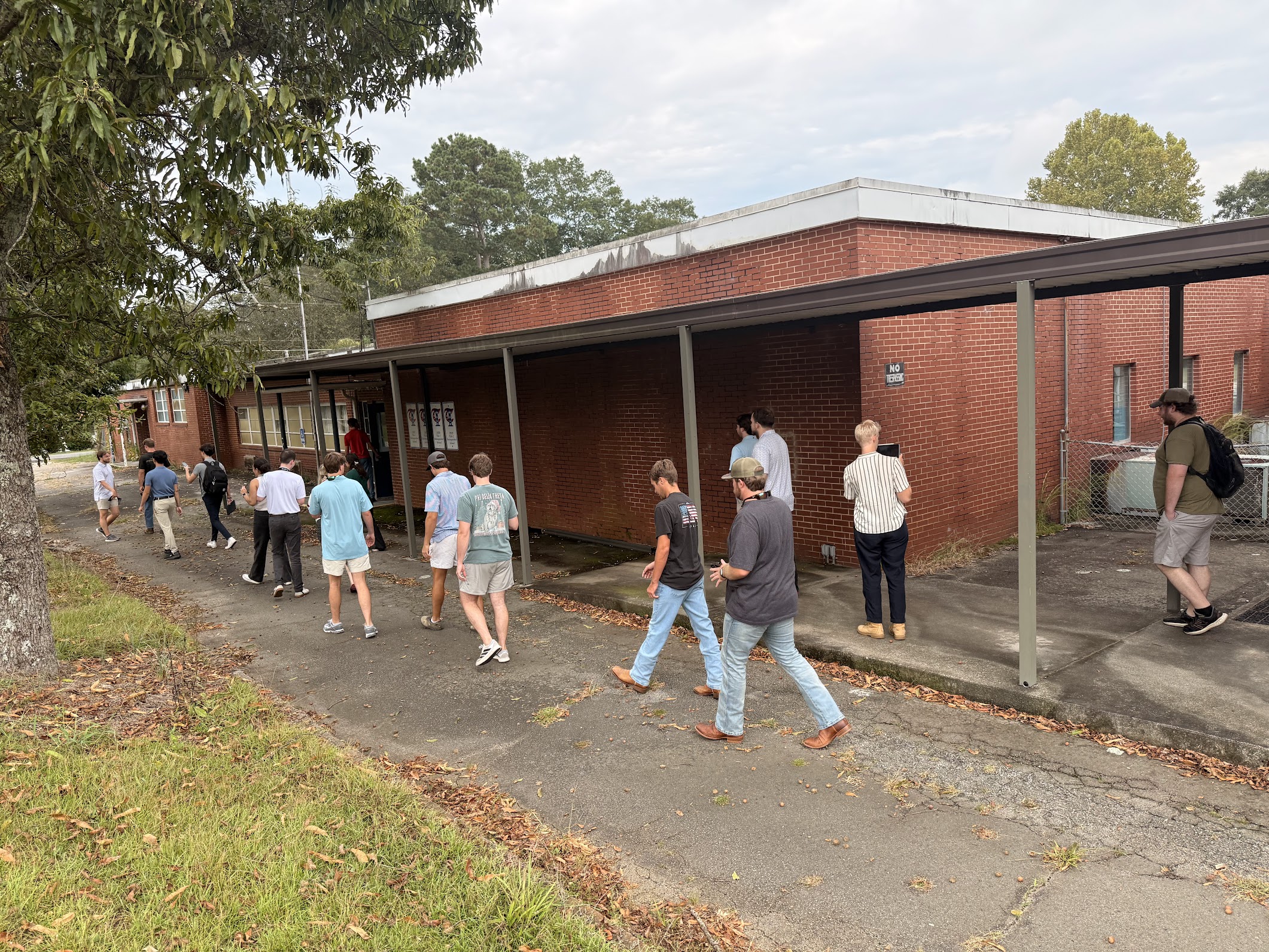 A group of students walks outside a brick building on a pavement sidewalk. The building and sidewalk are aging and the students are inspecting as a group, taking photos.