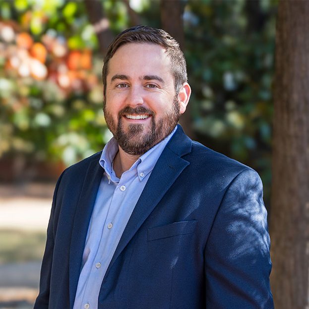 Matt Bilskie, wearing a blue suit, stands in front of a blurred background of trees