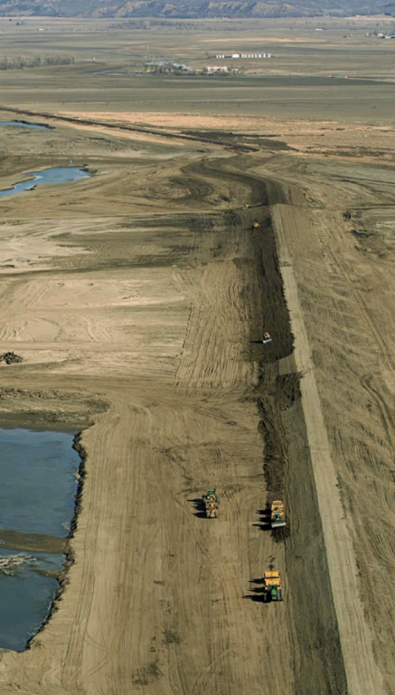 An aerial view of a levee setback under construction shows construction vehicles alongside an earthen levee to the right and a waterbody to the left.