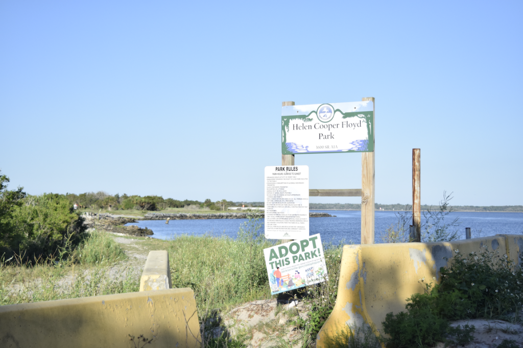 a tall park sign reading "Helen Cooper Floyd Park" in front of a stretch of rocky and green shoreline. Below the sign are other signs, reading "Park Rules" and "Adopt This Park"